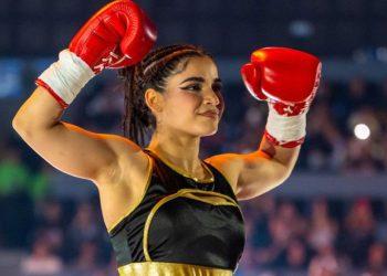 Female Boxer in Black and Gold Outfit Raises Red Gloves in Victory Pose Inside a Arena with Cheering Crowd Behind Her. • Guadalupe Informa