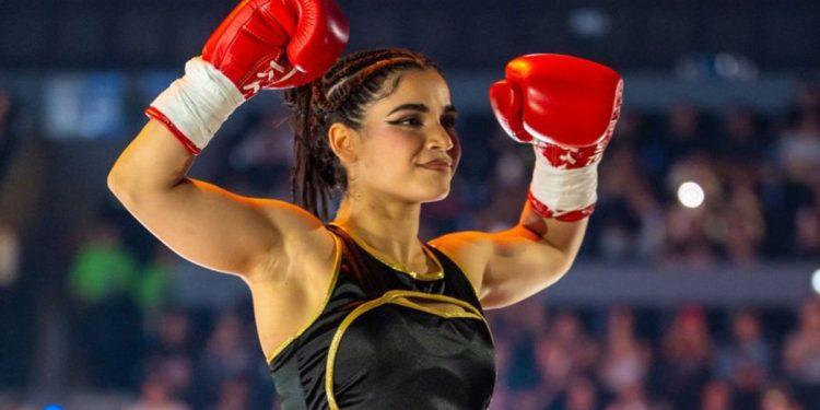 Female Boxer in Black and Gold Outfit Raises Red Gloves in Victory Pose Inside a Arena with Cheering Crowd Behind Her. • Guadalupe Informa