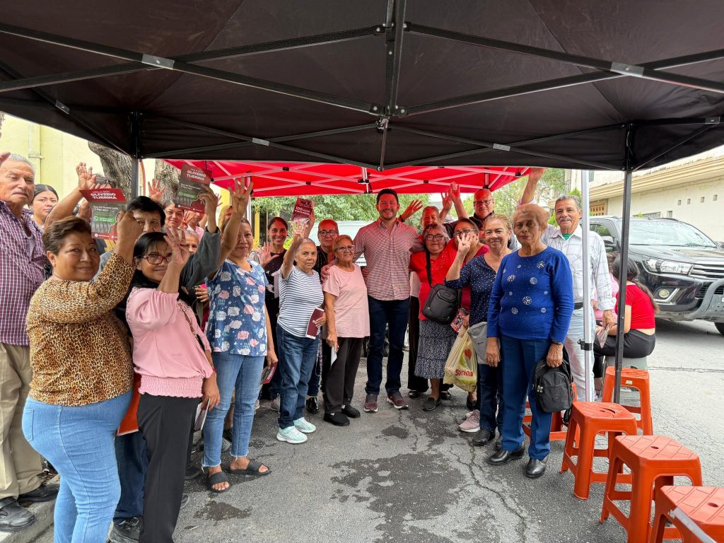 Diverse Group of People Under a Black and Red Canopy Outside, Smiling and Raising Their Hands for a Group Photo at a Community Event. • Guadalupe Informa