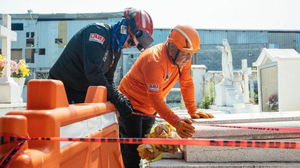 Two Workers in Helmets Lean over a Cemetery Grave, Placing Flowers Amid Orange Safety Tape and Barriers.6e14a74d6 the Scene Shows Tombstones in the Background. • Guadalupe Informa