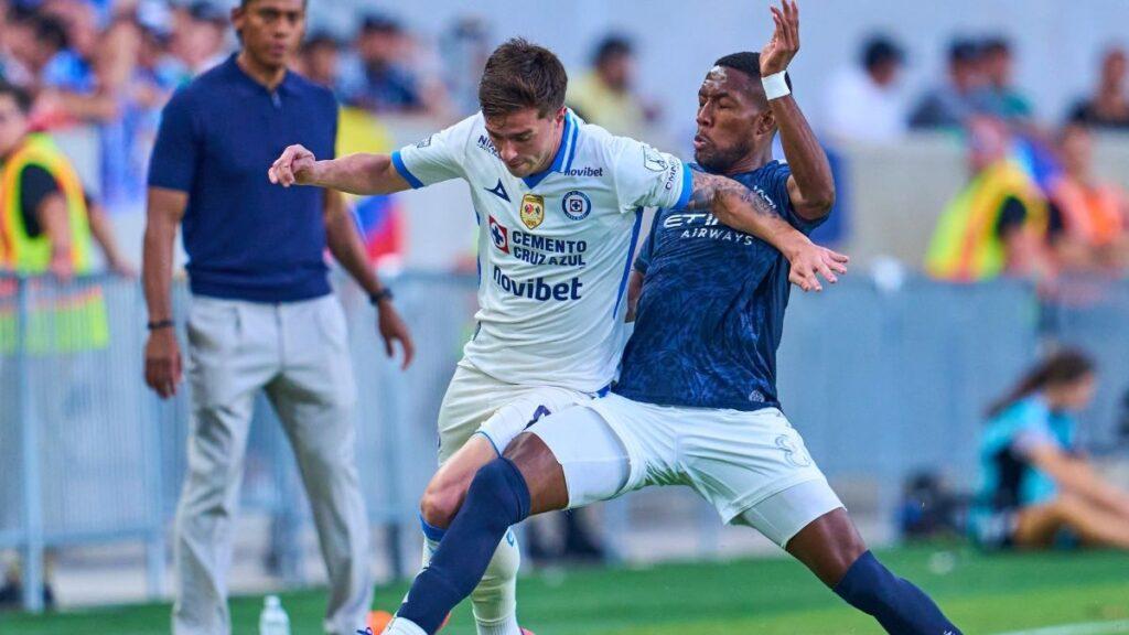 Two Soccer Players Contest the Ball During a Match, One in a White Jersey and the Other in a Dark Blue Kit, on a Green Field with Spectators in the Background. • Guadalupe Informa